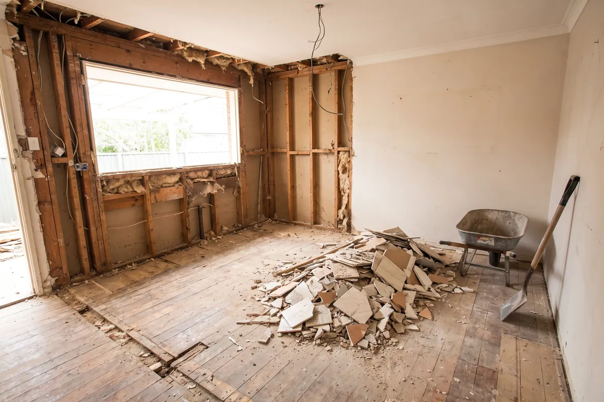 House interior mid strip-out with the old kitchen removed, exposed timber wall studs, rubble swept into a pile and a wheelbarrow standing by