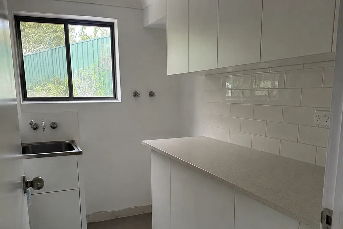 Completed ReachOut laundry renovation with white overhead and base cabinetry, a stone benchtop, subway tile splashback and a stainless tub